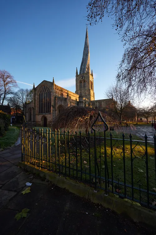 This architectural photograph, taken in the morning during the winter season, features the railings in the foreground of the Church of St Mary and All Saints in Chesterfield, Derbyshire, United Kingdom. The image showcases the iconic twisted spire of the church, also known as Chesterfield Parish Church, which is a well-known landmark in the area. The church building is surrounded by leafless trees, indicative of winter, while the early sunlight casts long shadows across the grounds. The architectural details of the church, including its stone construction and large arched windows, are prominent, and the setting captures the historical character of Chesterfield in Derbyshire.